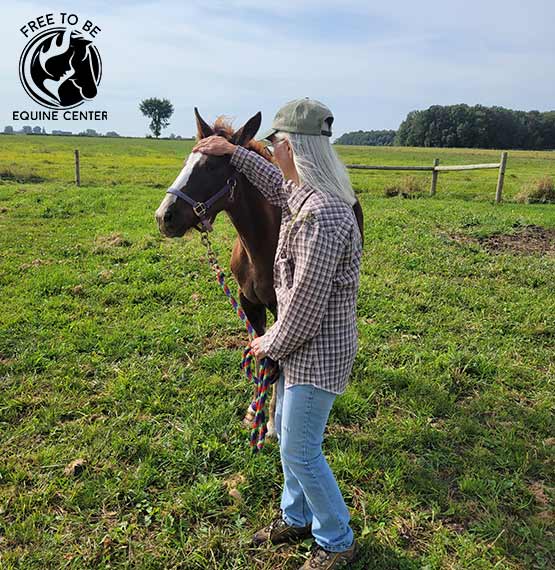 older woman holding the lead of a horse and petting the horse in a field with the Free to Be Equine Center logo in top left corner