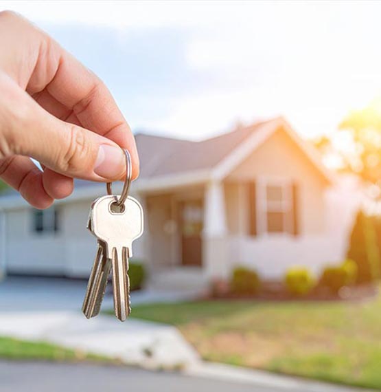 A hand is holding house keys in the foreground. In the background, softly blurred, is a small, manufactured or modular home during golden hour. 