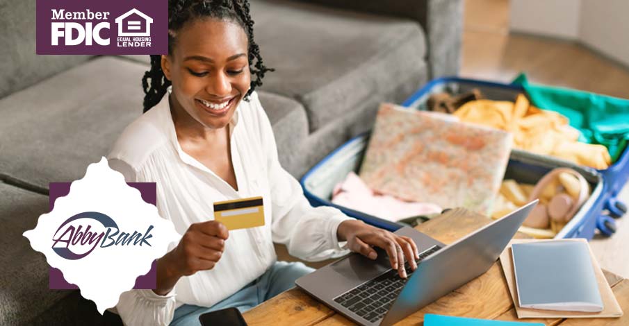 woman smiling with credit card while looking at laptop with open suitcase nearby