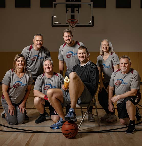 Brian Butch holding an ice cream cone while sitting in chair on a gym floor with basketball under one foot and AbbyBank team members around him