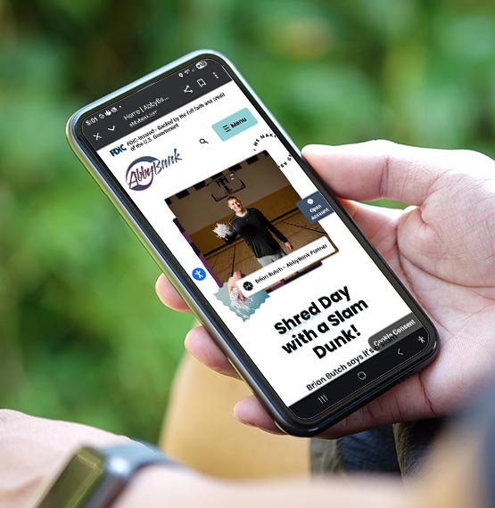 person on park bench looking at mobile phone with AbbyBank website pulled up showing Shred Day ad with Brian Butch